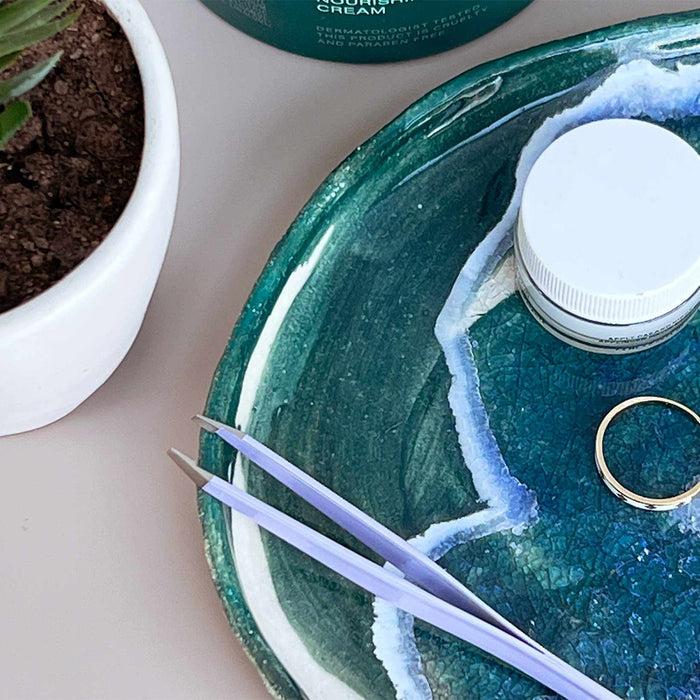 Flamingo Tweezers on a glazed teal ceramic dish that also holds a gold ring and a small jar of cosmetics